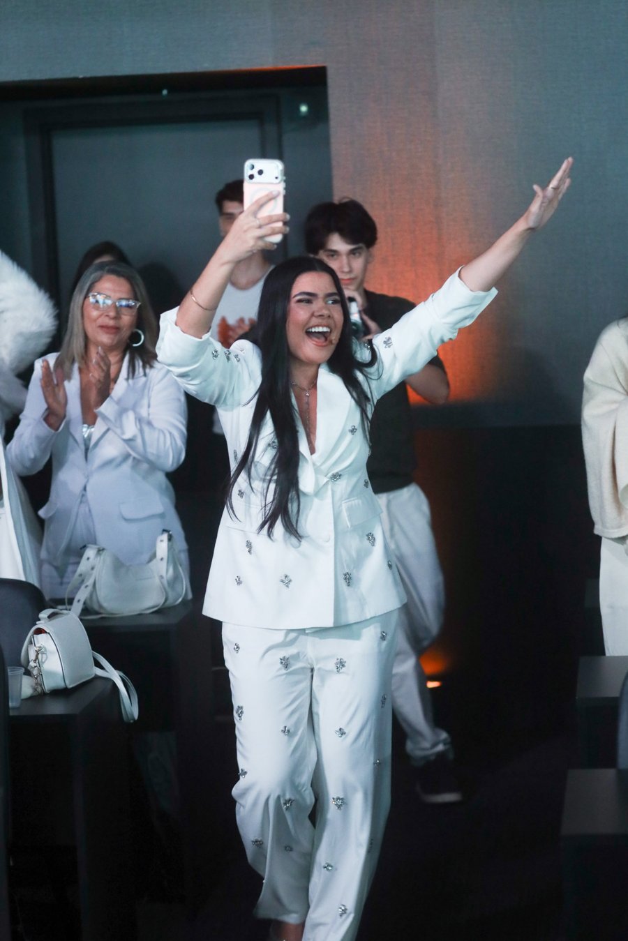 Andressa Diniz no palco, de terno branco, acenando para o público